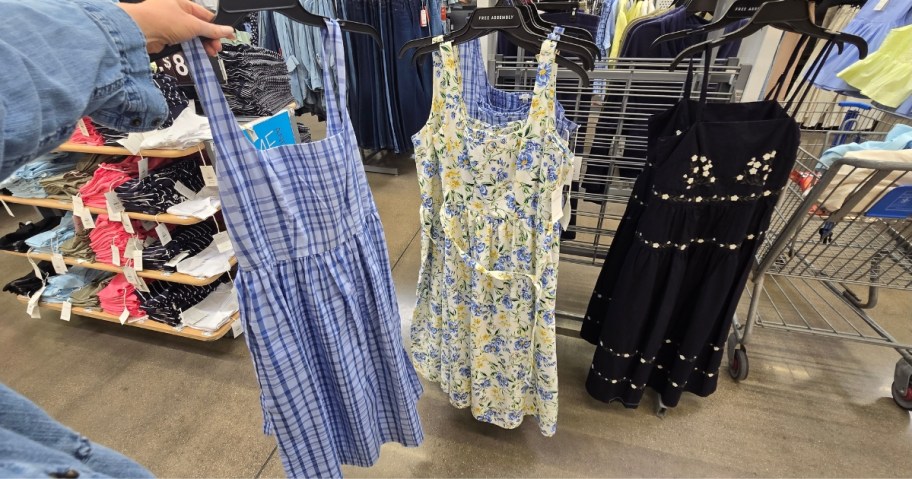 woman holding a blue dress on a hanger, a floral dress and black dress next to it