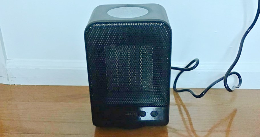 A black portable heater with a mesh front sits on a wooden floor, plugged into a white wall.