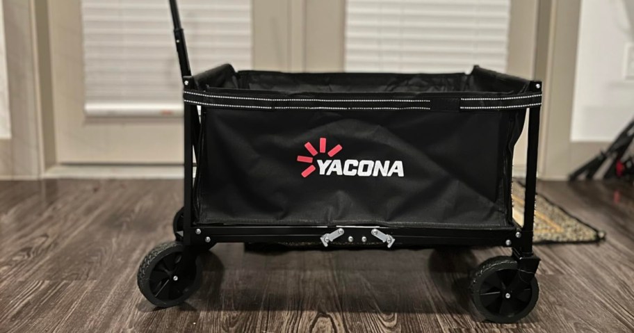 A black Yacona wagon with four wheels stands on a wooden floor indoors. It has a metal handle, white logo, and appears sturdy and functional.