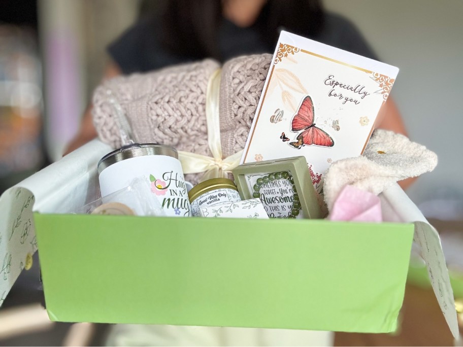woman holding a gift box with a blanket, mug, socks, candle, and card