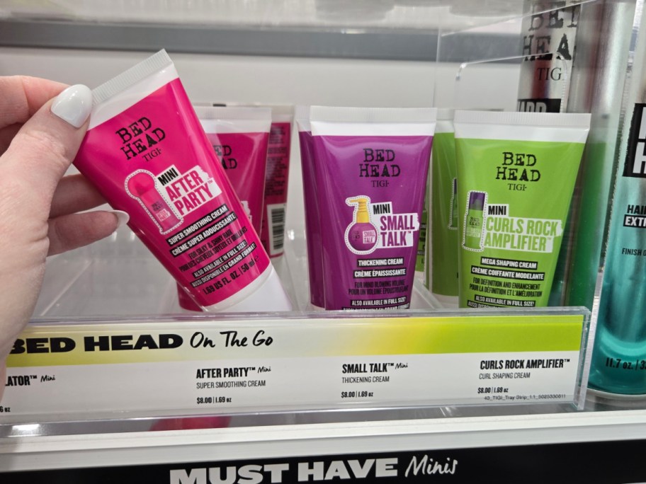 A hand holding a pink tube of "Bed Head Mini After Party" smoothing cream. Beside it are "Mini Small Talk" and "Curls Rock Amplifier" creams on a shelf.