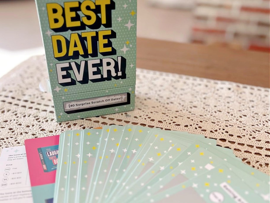 Green box reading "Best Date Ever!" with stars, showcasing a card deck for "40 Surprise Scratch Off Dates." Cards are spread on a lace tablecloth.