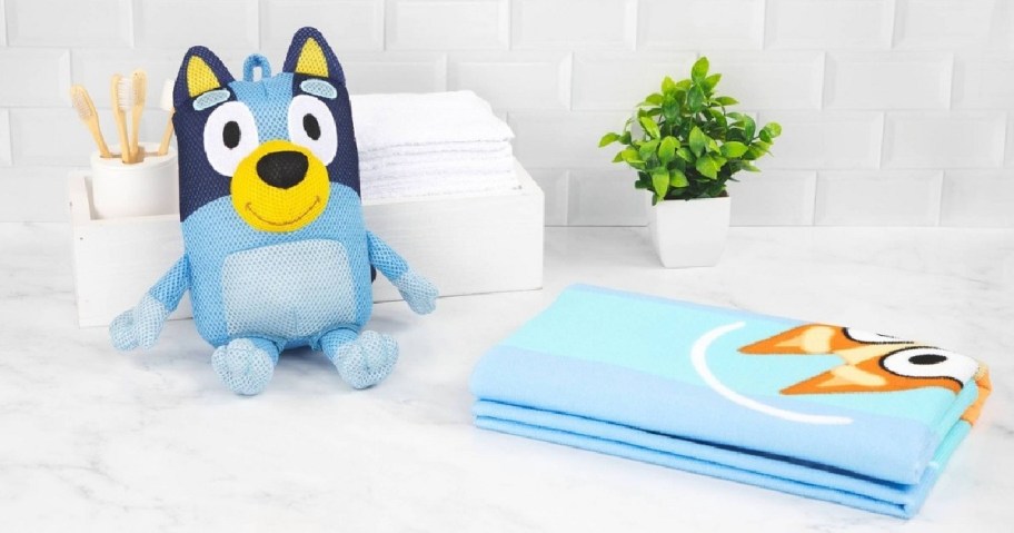 A plush toy resembling Bluey sits on a marble surface. Nearby is a folded matching Bluey towel, toothbrushes in a holder, and a small green plant.
