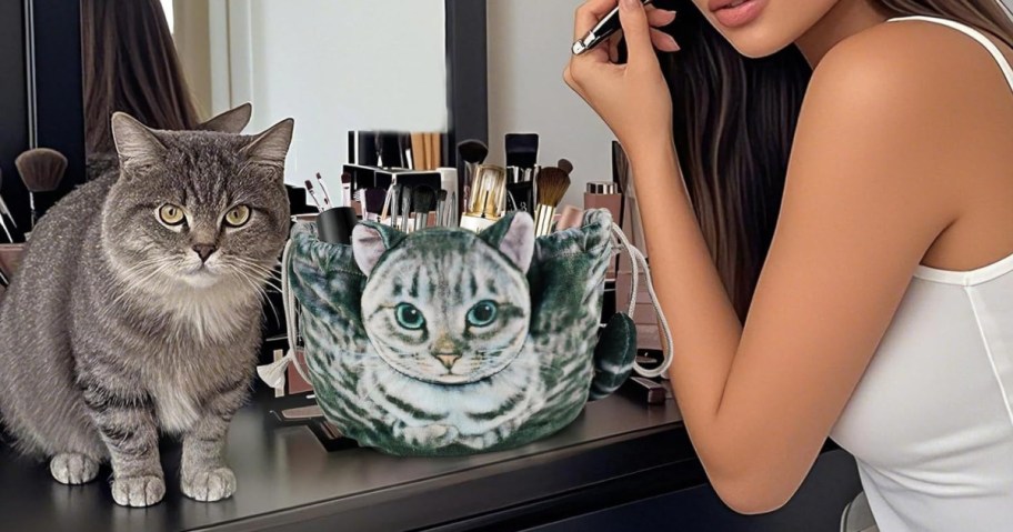 A gray cat sits on a vanity next to a bag printed with a cat face. A woman in a white tank top applies makeup.