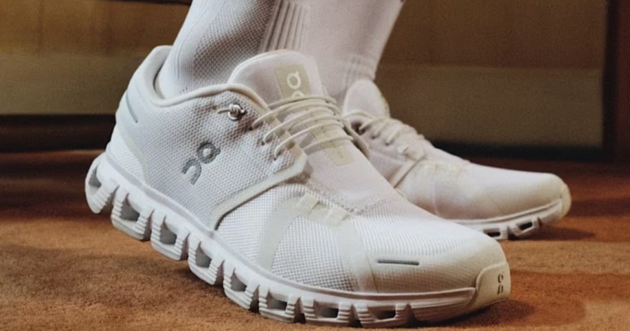 White running shoes with distinctive textured soles are worn with matching high socks on a brown carpet.