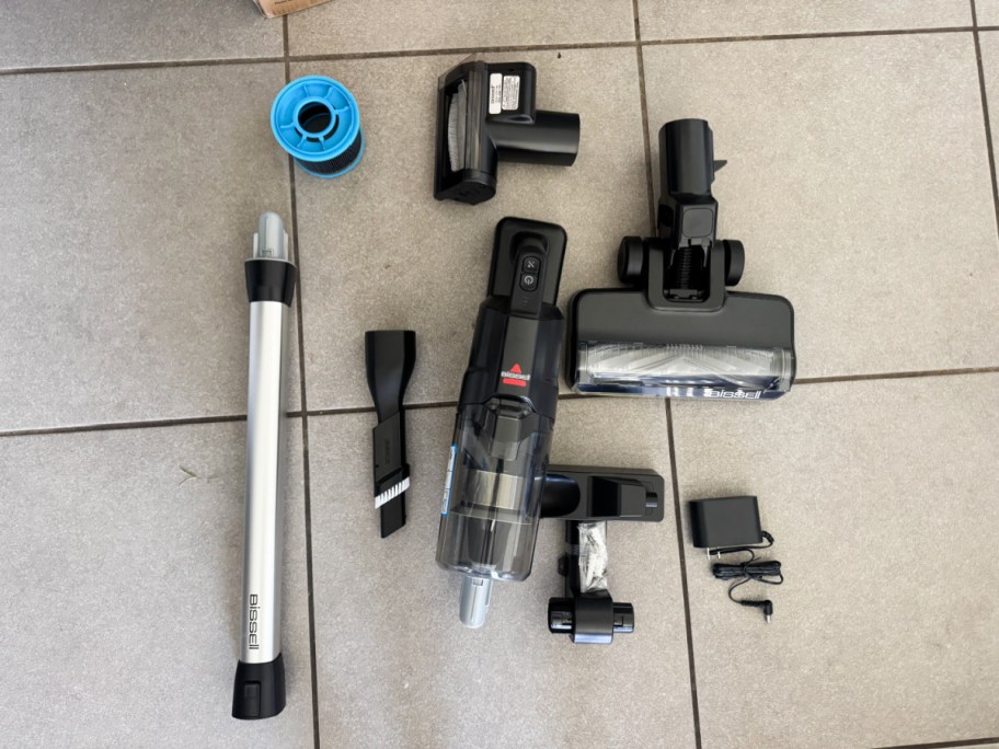 Vacuum cleaner parts are spread out on a tiled floor, including a main unit, brush heads, a tube, filter, adapter, and small tools.