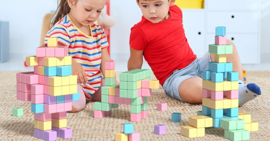 Two children sitting on a carpet play with colorful building blocks, creating structures.