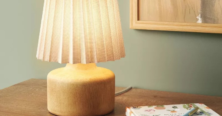 A warm, glowing table lamp with a pleated shade sits on a wooden desk next to a floral notebook and pen.