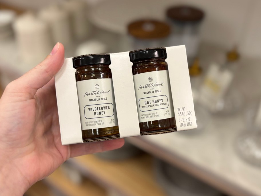 A hand holds two jars of Hearth & Hand honey in a white cardboard holder. The jars are labeled "Wildflower Honey" and "Hot Honey." The background is blurred.