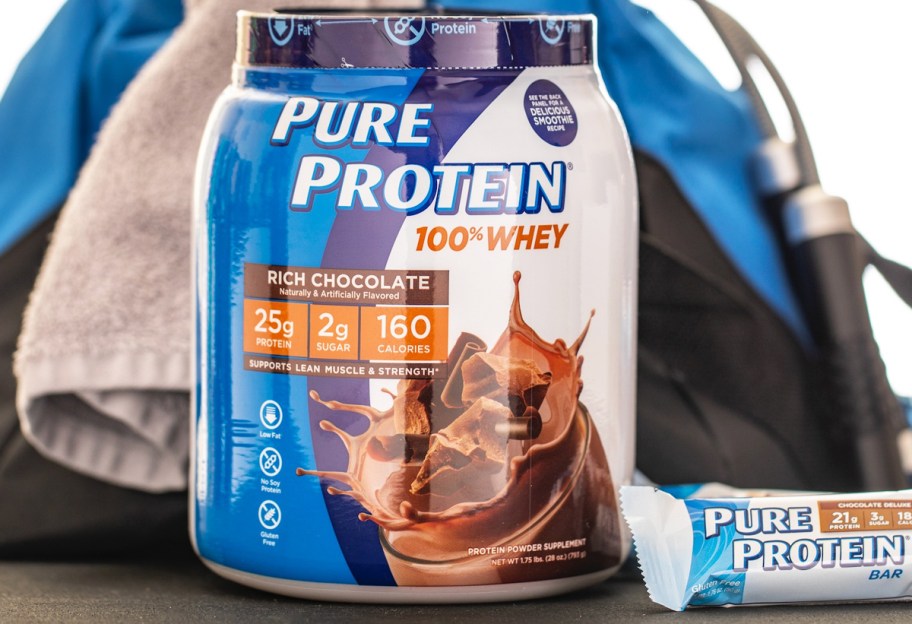 container of Pure Protein Rich Chocolate Protein Powder and protein bar in front of gym bag and towel