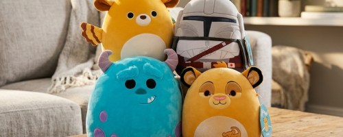 Four plush toys sit on a wooden table in a cozy living room. They resemble a deer, a helmeted figure, a blue creature with horns, and a lion cub.