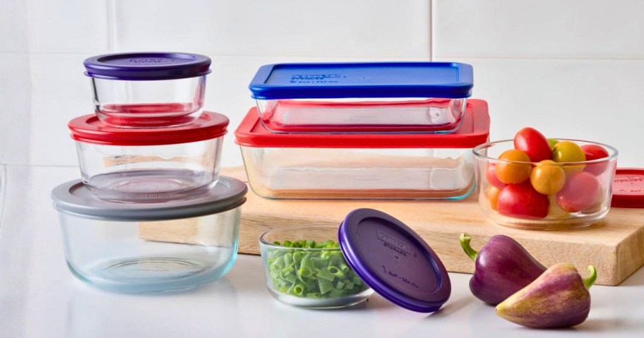 a set of round and rectangular Pyrex glass storage containers with lids with food around them