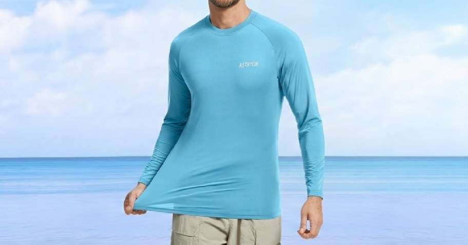 A man wearing a light blue long-sleeve top stands on a beach, stretching the fabric.