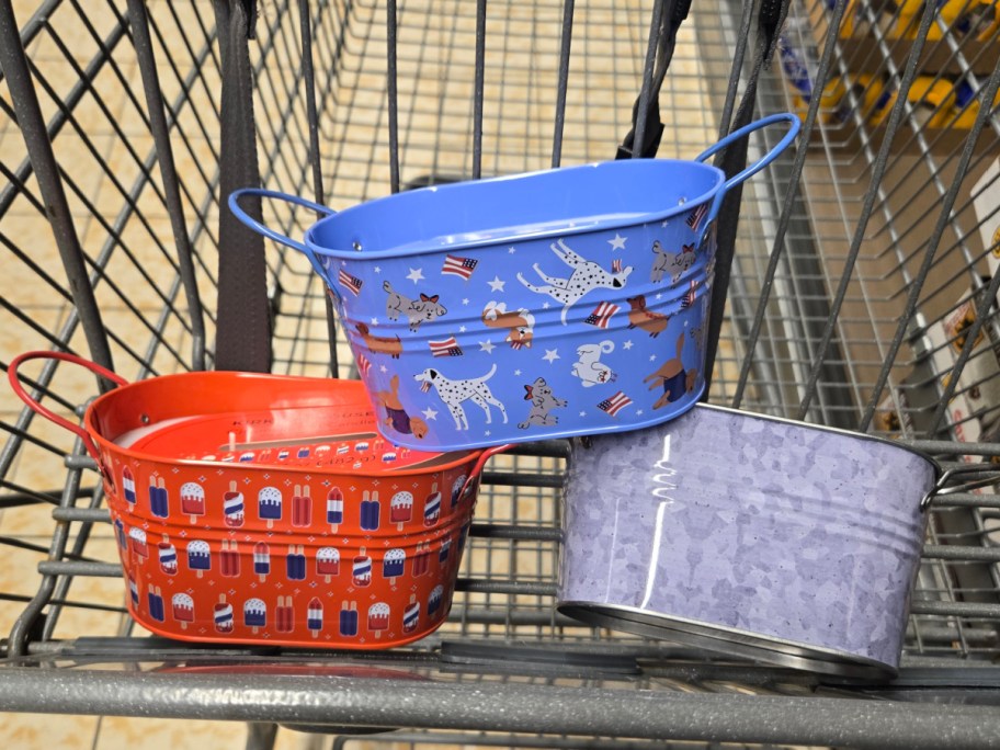 Three decorative metal citronella candles are placed in a shopping cart. The red candle has fireworks designs, the blue one features dog patterns, and the gray one is textured.
