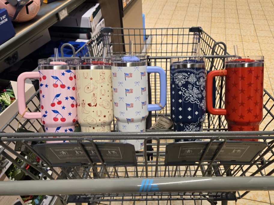 Shopping cart containing five large, colorful travel mugs with different patterns: cherries, western, flags, bandana, and stars.